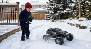 A young Canadian boy in warm winter clothing operating a rugged, all-terrain remote controlled monster truck through deep snow in a backyard.